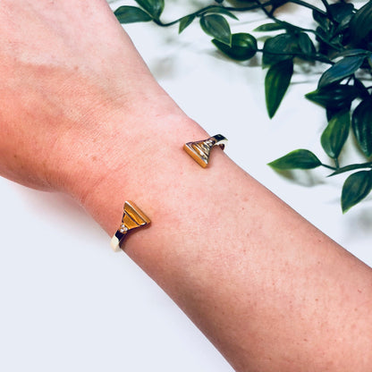 Gold toned triangle cuff bracelet on a person's wrist next to green leaves