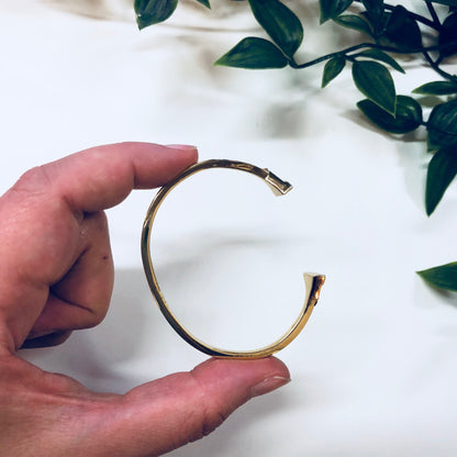 Gold toned open cuff bracelet with triangle shape held in hands near leaves