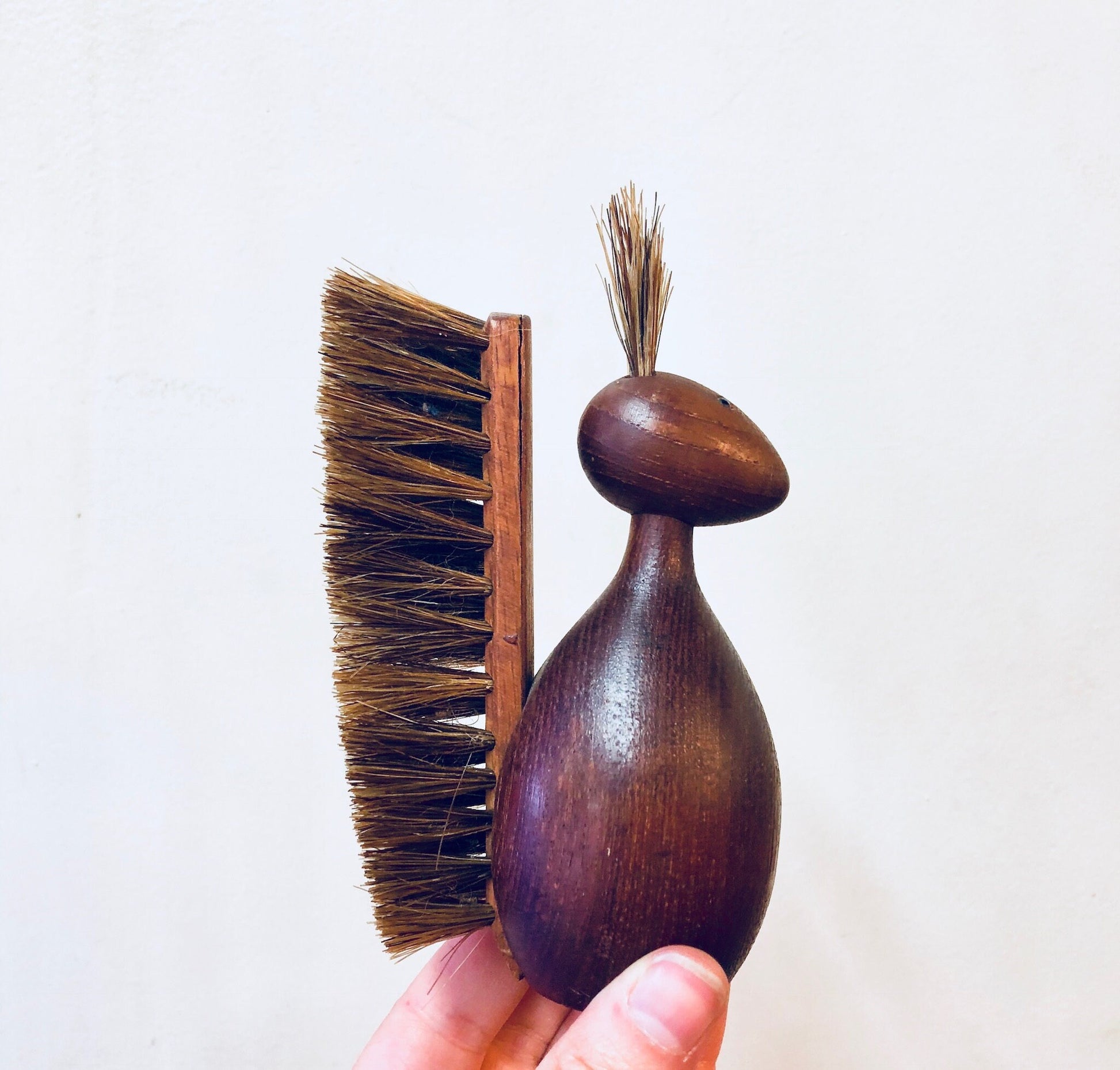 Antique wooden brush shaped like an animal, with bristles on the body and tail, held in a person's hand against a plain white background.