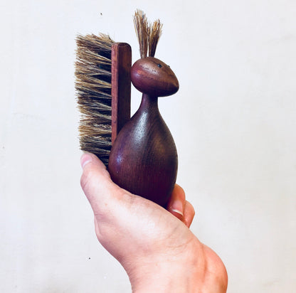 Hand holding a vintage wooden brush with bristles and a carved animal shape, likely a squirrel, against a plain background.