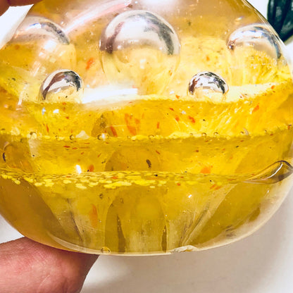 Close-up of a yellow glass paperweight with colorful speckled design featuring orange accents, held in hand against a white background, ideal for vintage office decor.