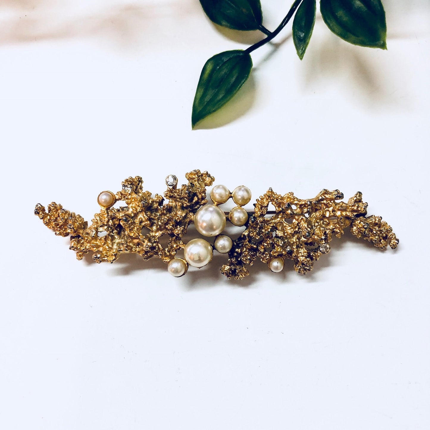 Vintage gold-toned brooch with faux pearls and intricate gold-toned branches, displayed next to green leaves on a white background, showcasing a statement piece of costume jewelry.