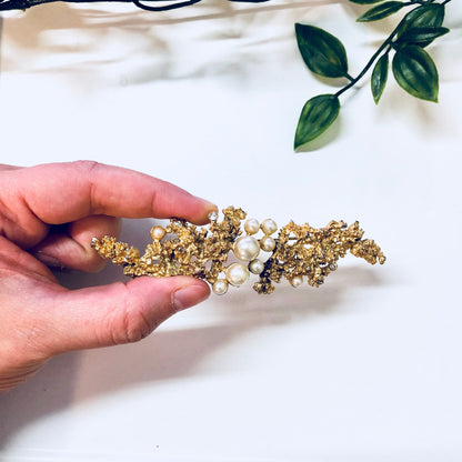 Vintage gold-toned brooch with faux pearls held in hand against white fabric with green leaves in background