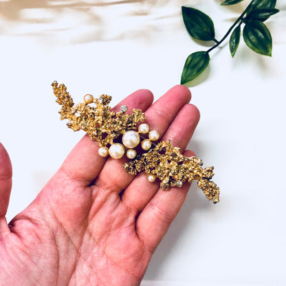 Vintage gold-toned brooch with faux pearls and intricate leaf design, held in palm of hand against white fabric background with green leaves.