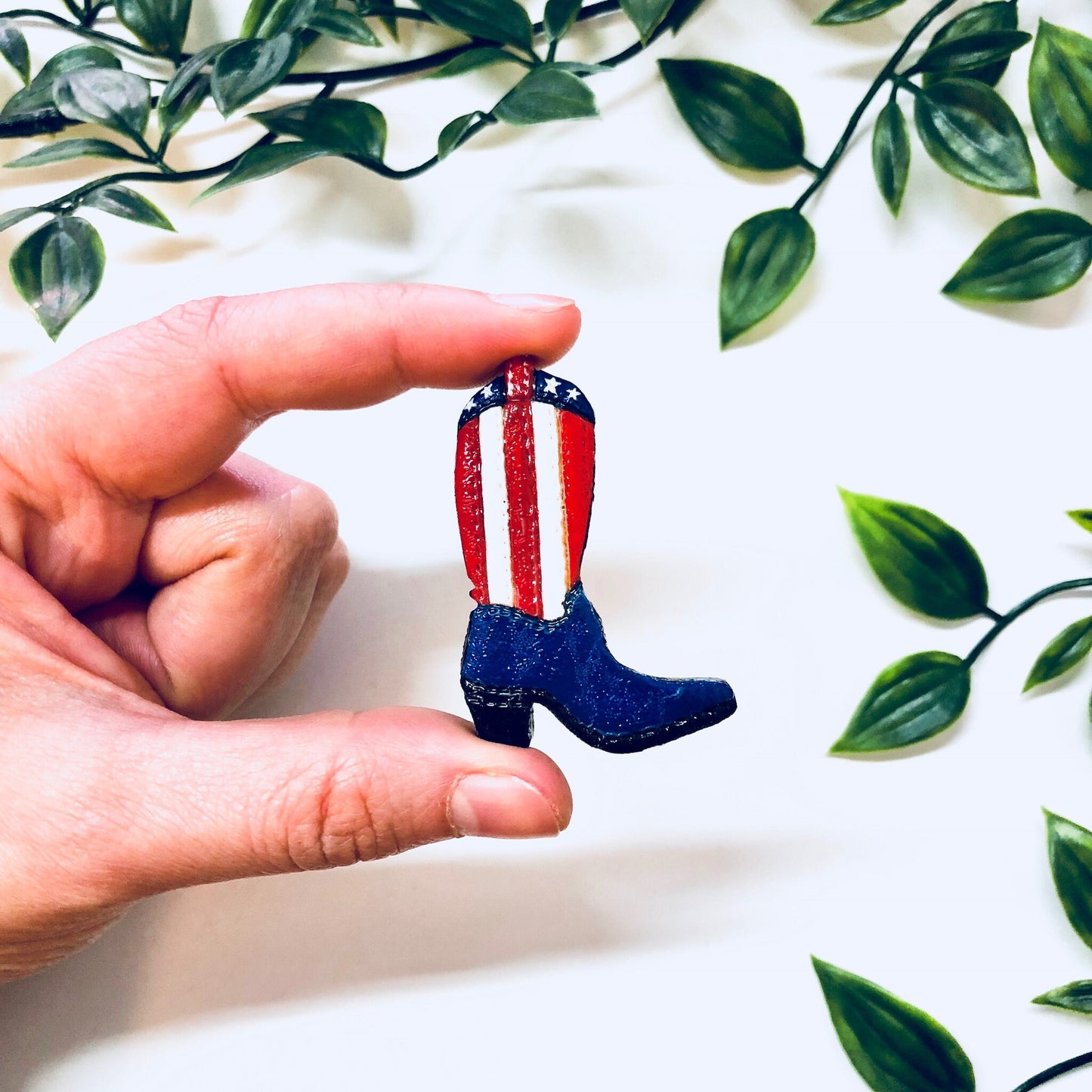 Vintage American flag cowboy boot pin held in front of green foliage background