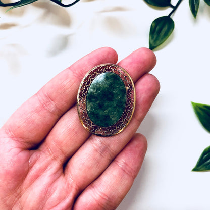 Vintage 12K gold filled brooch with green and white stone in an ornate gold setting, held in a person's hand with green leaves in the background.