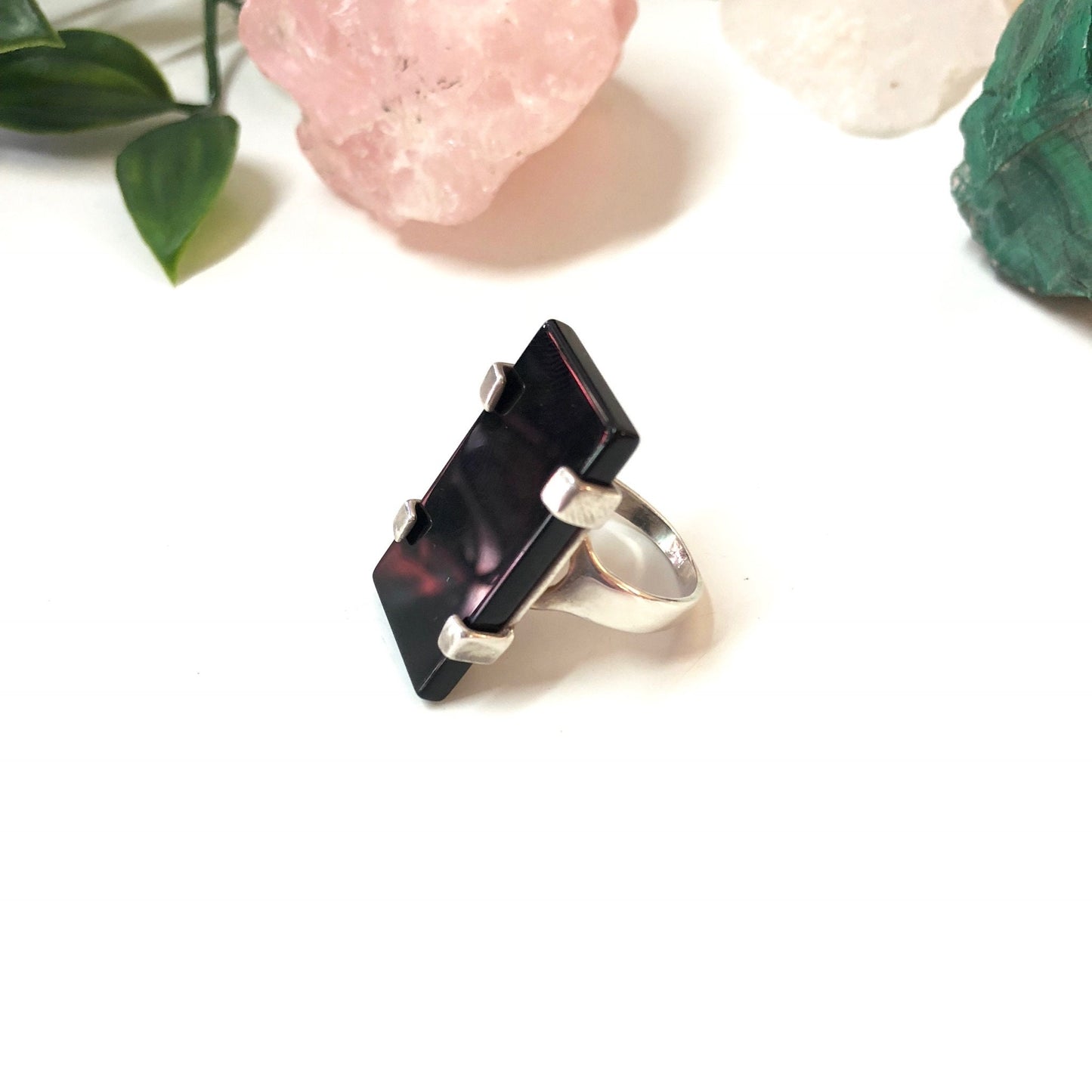 Vintage sterling silver ring featuring a black onyx stone in a minimalist rectangular setting, displayed against a white background with rose quartz and green leaves, capturing the elegance and timeless appeal of this unique vintage jewelry piece.