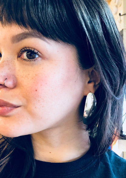 The image shows a close-up portrait of a young woman with dark hair and bangs, wearing large silver hoop earrings. She has freckles on her cheeks and is looking off to the side with a slight smile. The earrings are a prominent feature, appearing sleek and shiny against her dark hair.