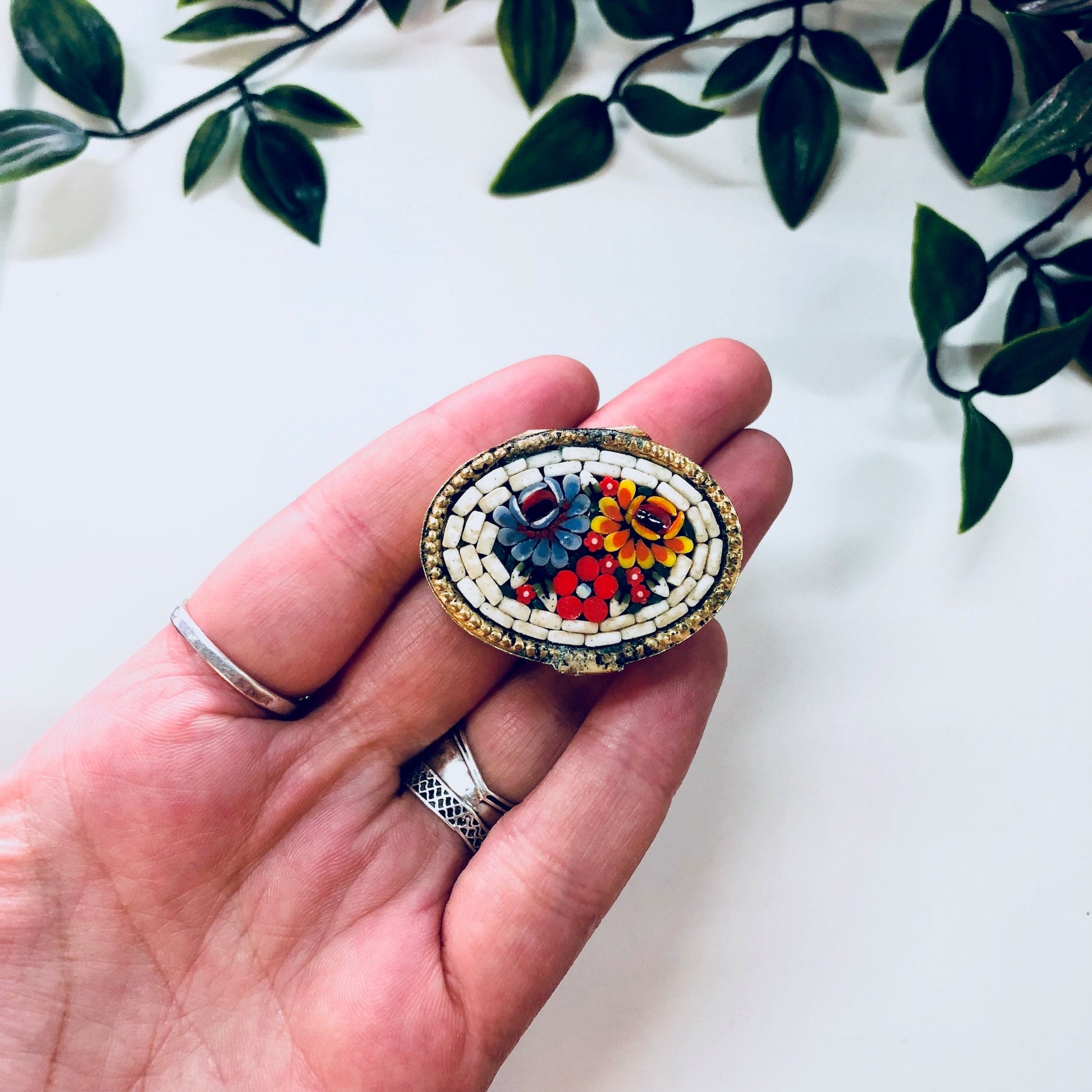 Vintage millefiori mosaic pill box held in hand against white background with green leaves, gold toned trinket case with red, blue, orange and white swirl paisley design.