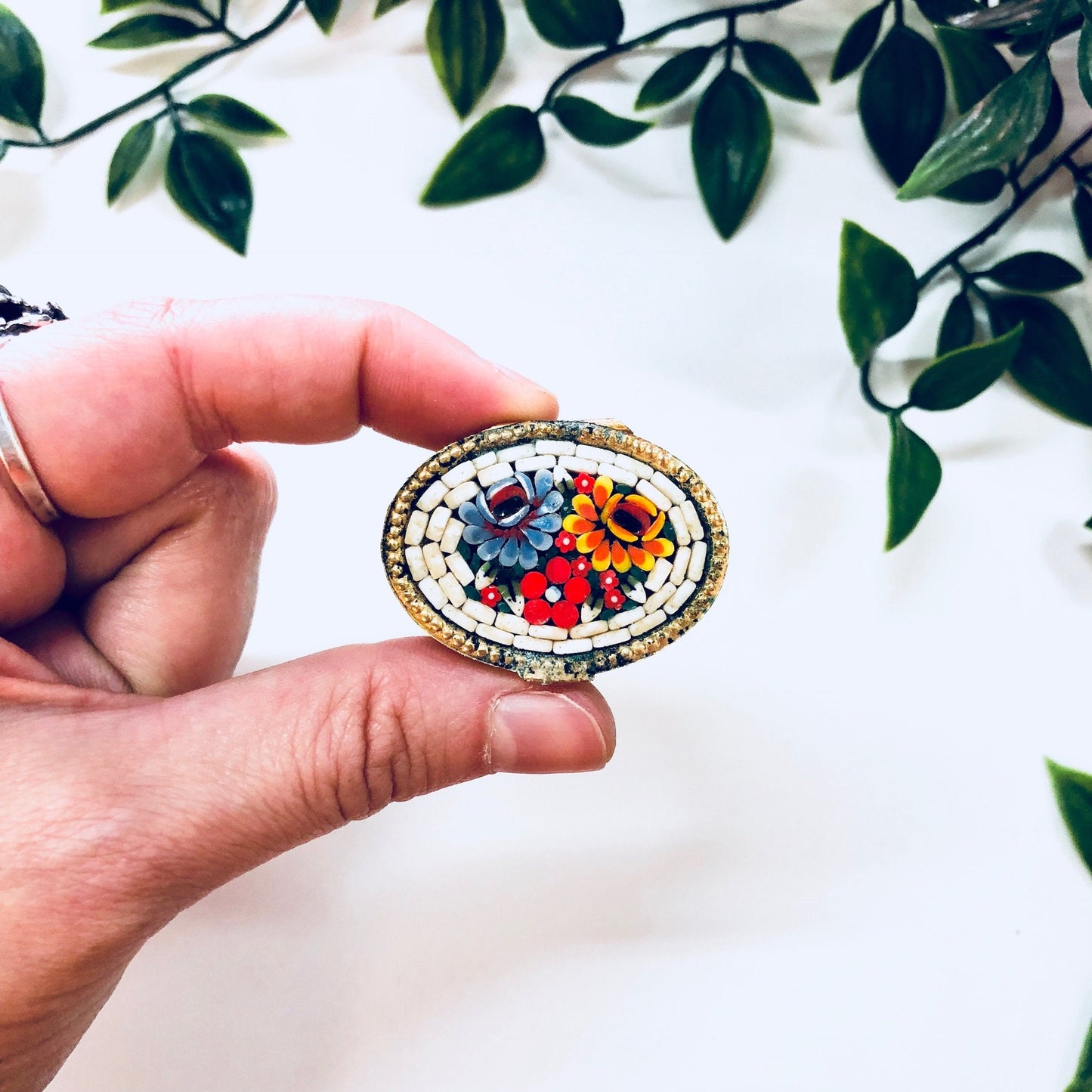 Hand holding a vintage gold-toned oval trinket box featuring a colorful millefiori mosaic design in red, blue, orange and white, surrounded by green leaves.