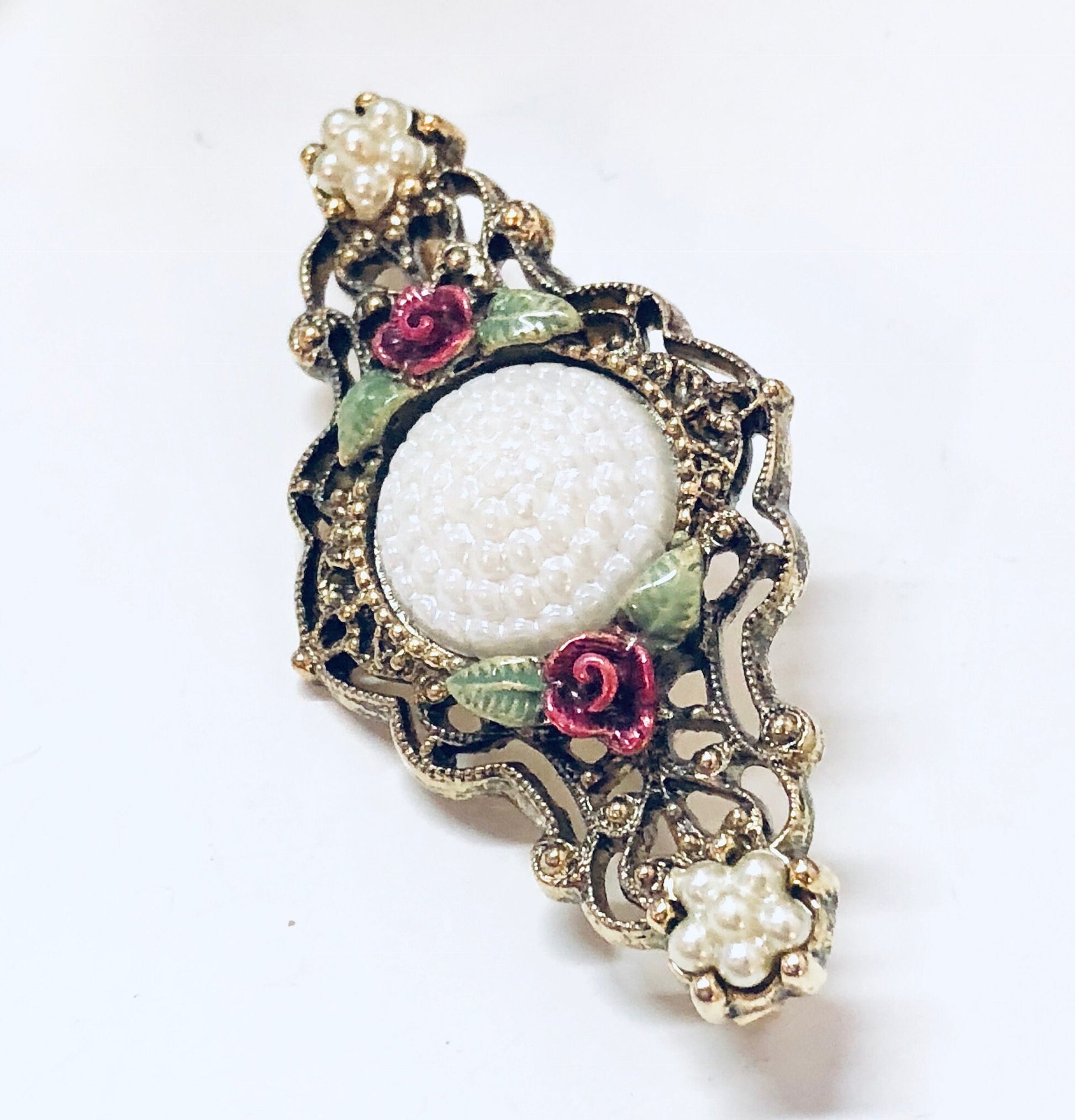 Ornate vintage gold-toned brooch with intricate floral design featuring white beads and red and green enamel accents, resembling costume jewelry from a bygone era.