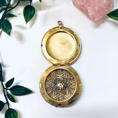 Vintage gold toned round locket pendant with intricate engraved design, featuring leaves and swirls, displayed on white cloth next to pink quartz and green leaves.