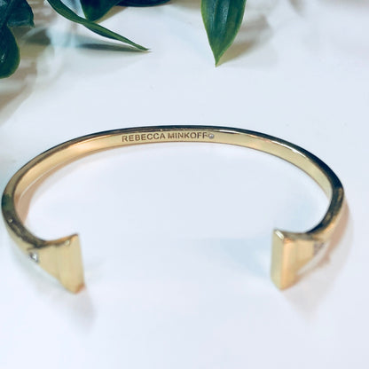 Gold toned Rebecca Minkoff cuff bracelet with triangle shapes on white marble surface next to green leaves