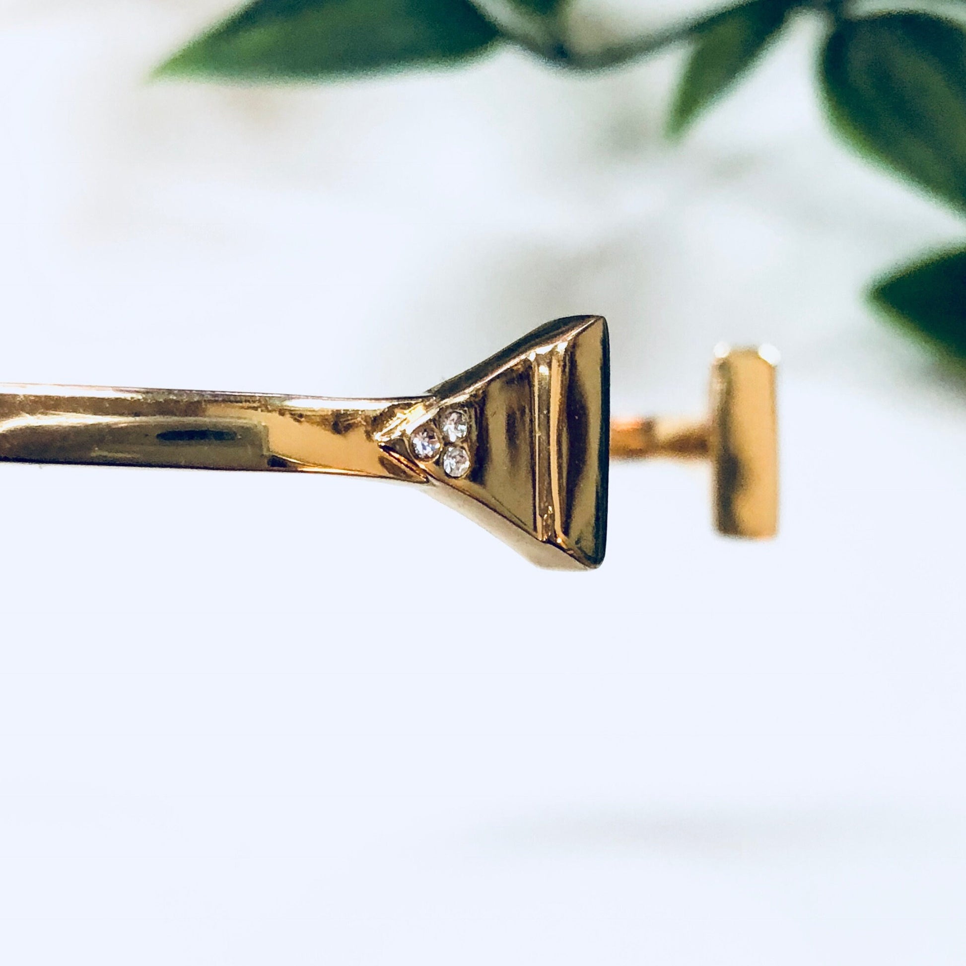 Gold toned Rebecca Minkoff cuff bracelet with triangle shape and diamond accents, photographed on a white background with green foliage.