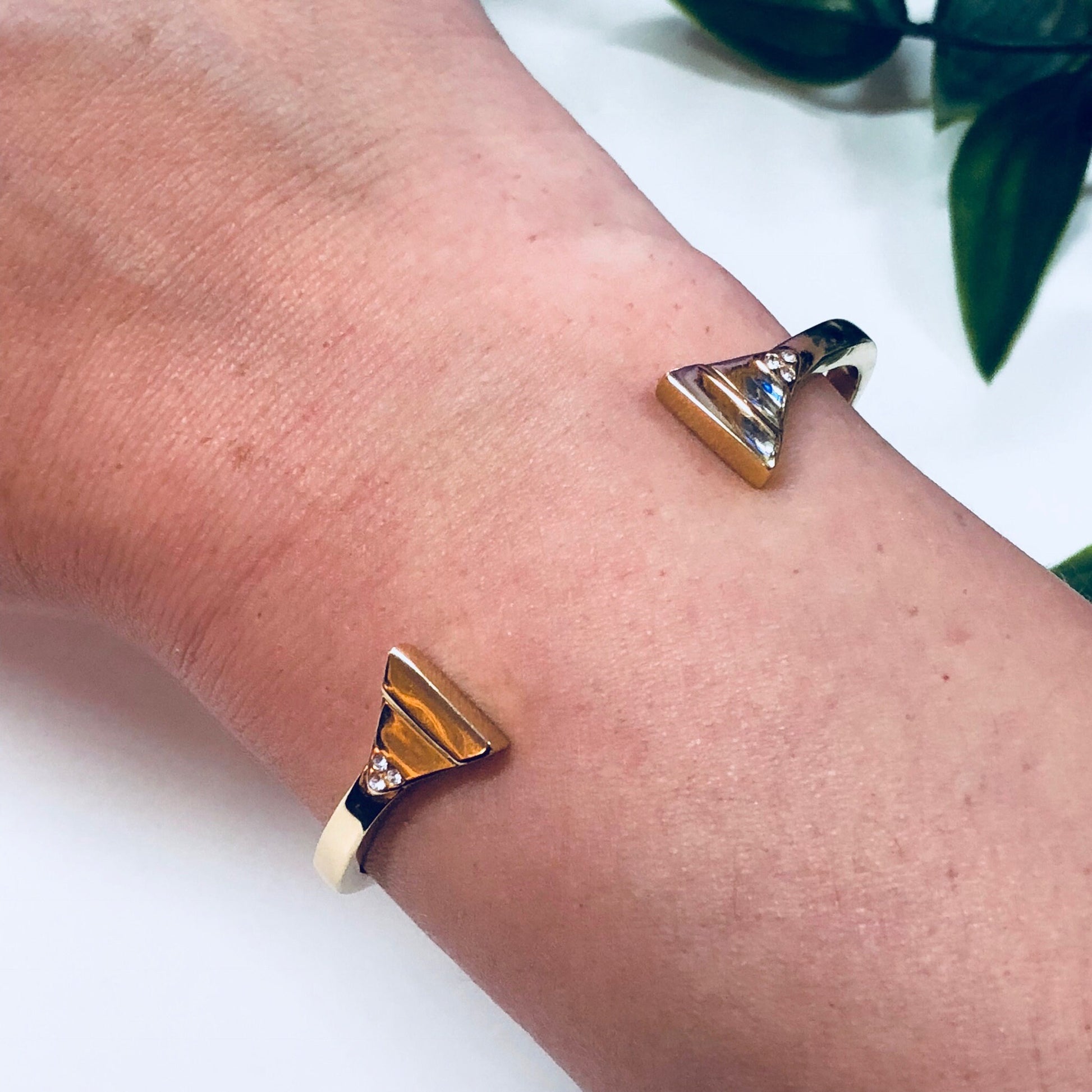 Gold toned triangle-shaped Rebecca Minkoff cuff bracelet on a person's wrist next to green leaves