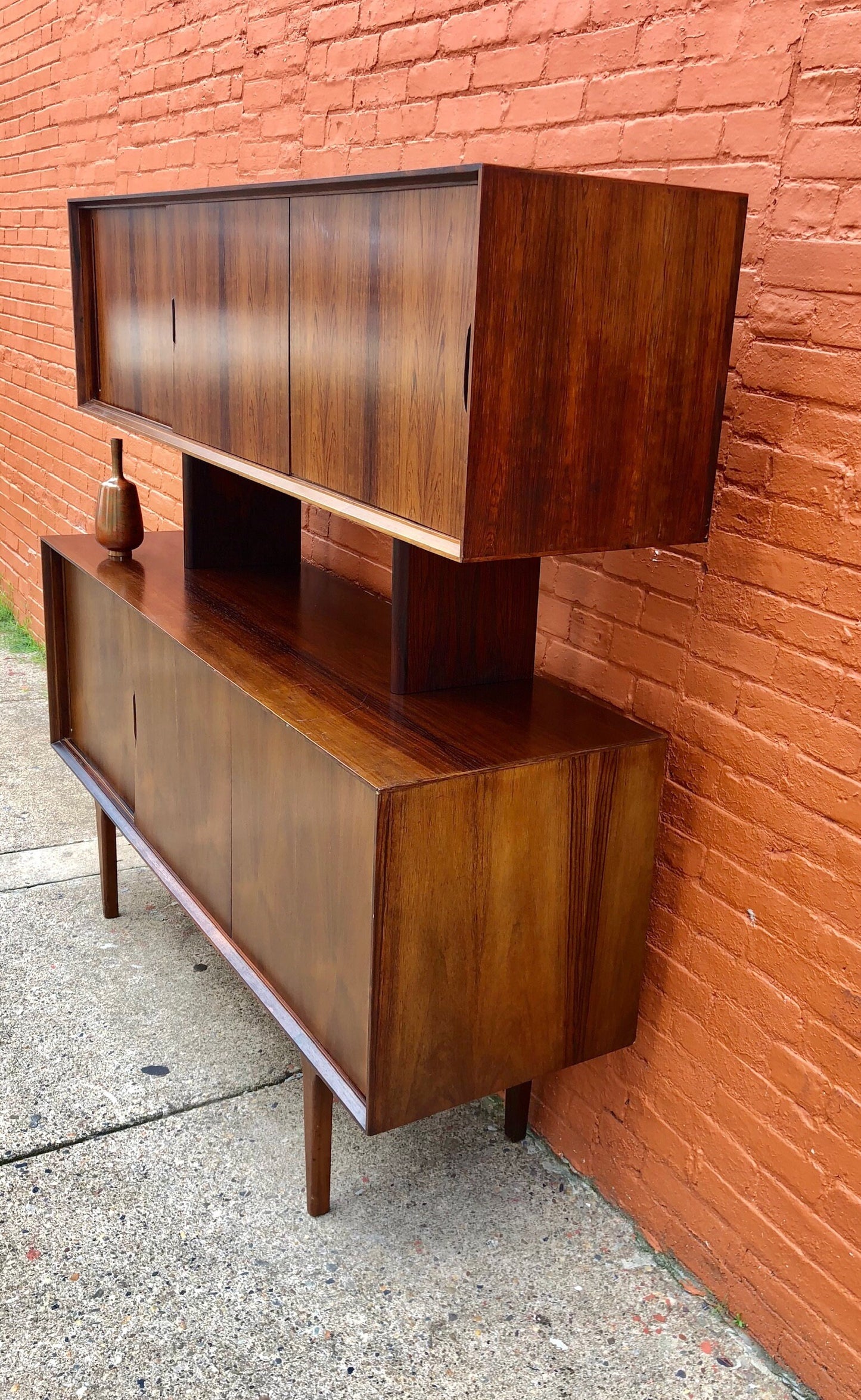 Elegant mid-century modern teak wood credenza buffet against a brick wall backdrop, featuring sleek lines, sliding cabinet doors and open shelving in a rich warm wood finish.