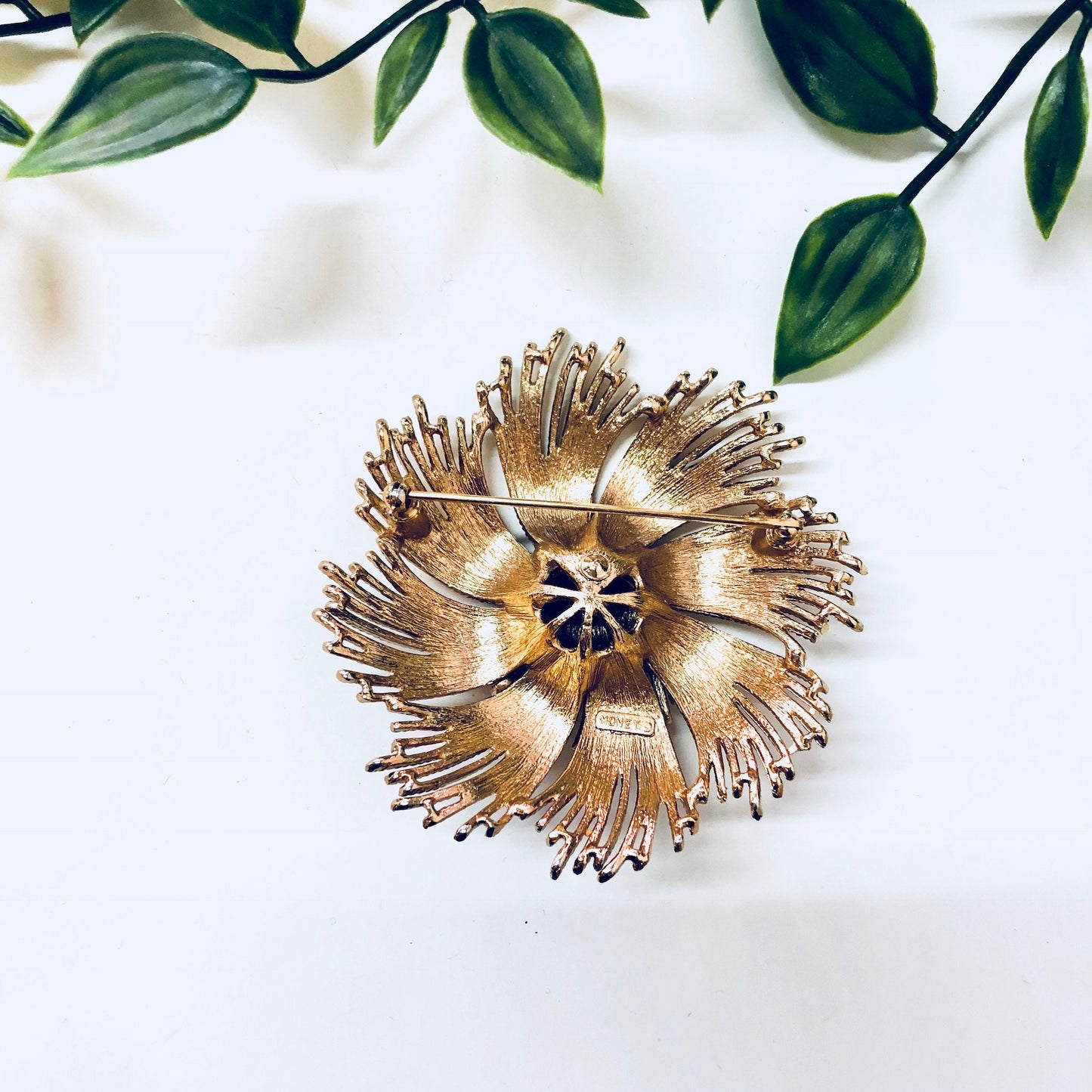 Vintage gold-toned swirl brooch by Monet featuring a unique floral design with blue stone center, displayed on a white background with green foliage.