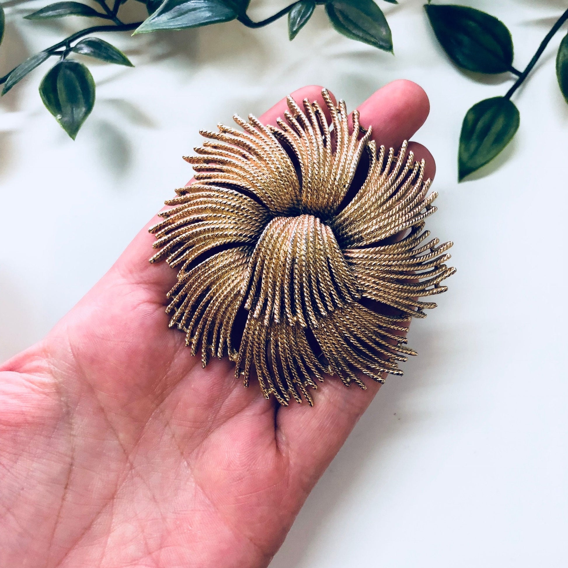 Vintage gold-toned swirl brooch by Monet, held in hand against white fabric with green leaves in background, statement jewelry piece with intricate textured design resembling a fireworks burst or sea anemone.