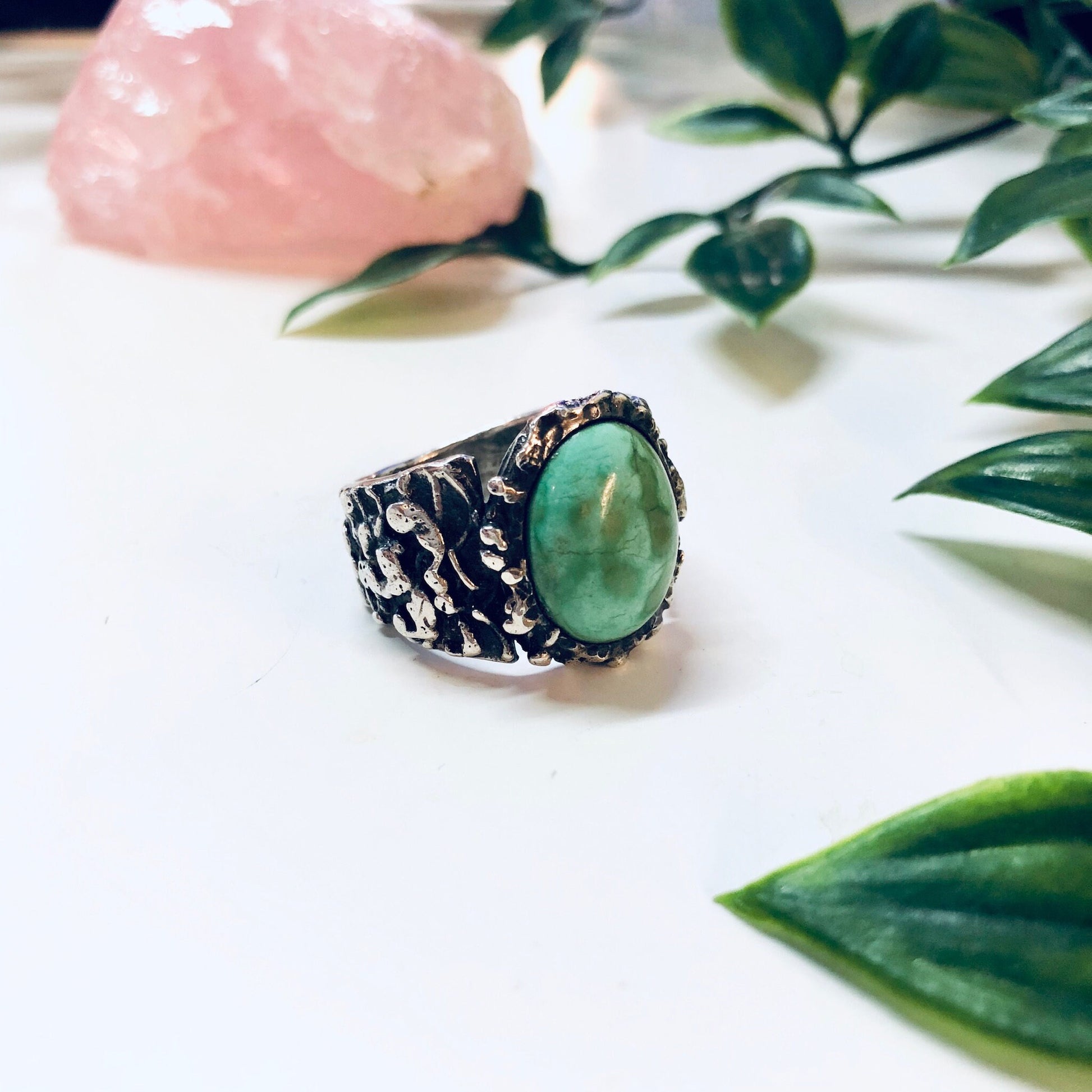 Vintage silver ring with an oval turquoise stone set in an ornate, textured sterling silver band, photographed with green foliage and a rose quartz crystal.