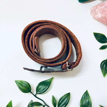 Vintage brown leather cowboy belt with hook buckle on white background surrounded by green leaves