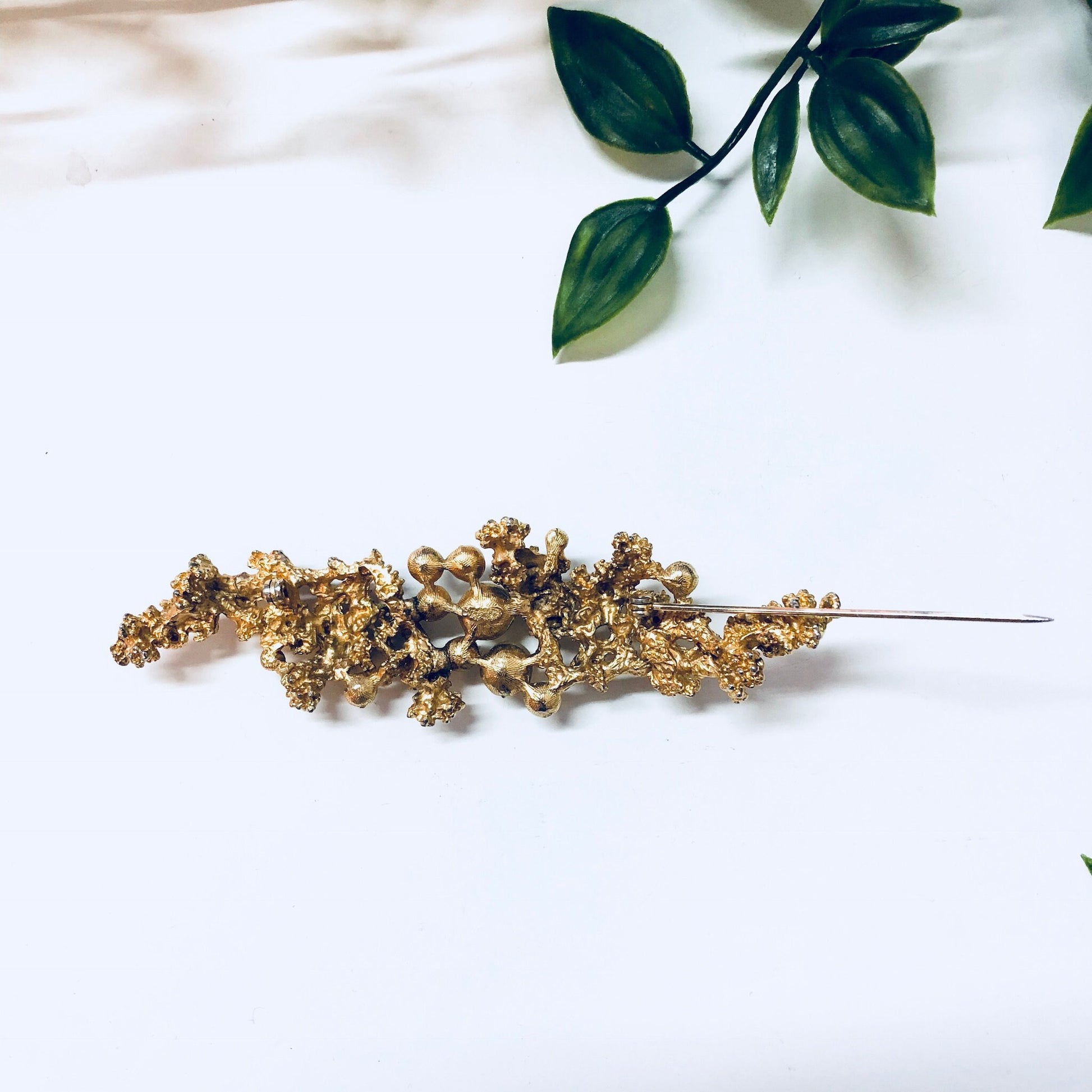 Vintage gold-toned brooch with faux pearls and intricate design, placed on white fabric with green leaves in the background, creating an elegant statement jewelry piece.