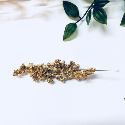 Vintage gold-toned brooch with faux pearls and intricate design, placed on white fabric with green leaves in the background, creating an elegant statement jewelry piece.