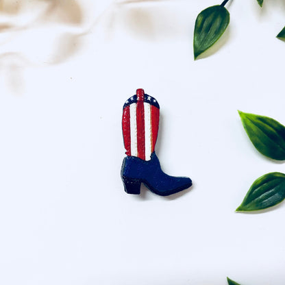 Vintage enamel pin in the shape of a blue cowboy boot with red and white vertical stripes and stars, resembling the American flag, displayed with green leaves on a white background.