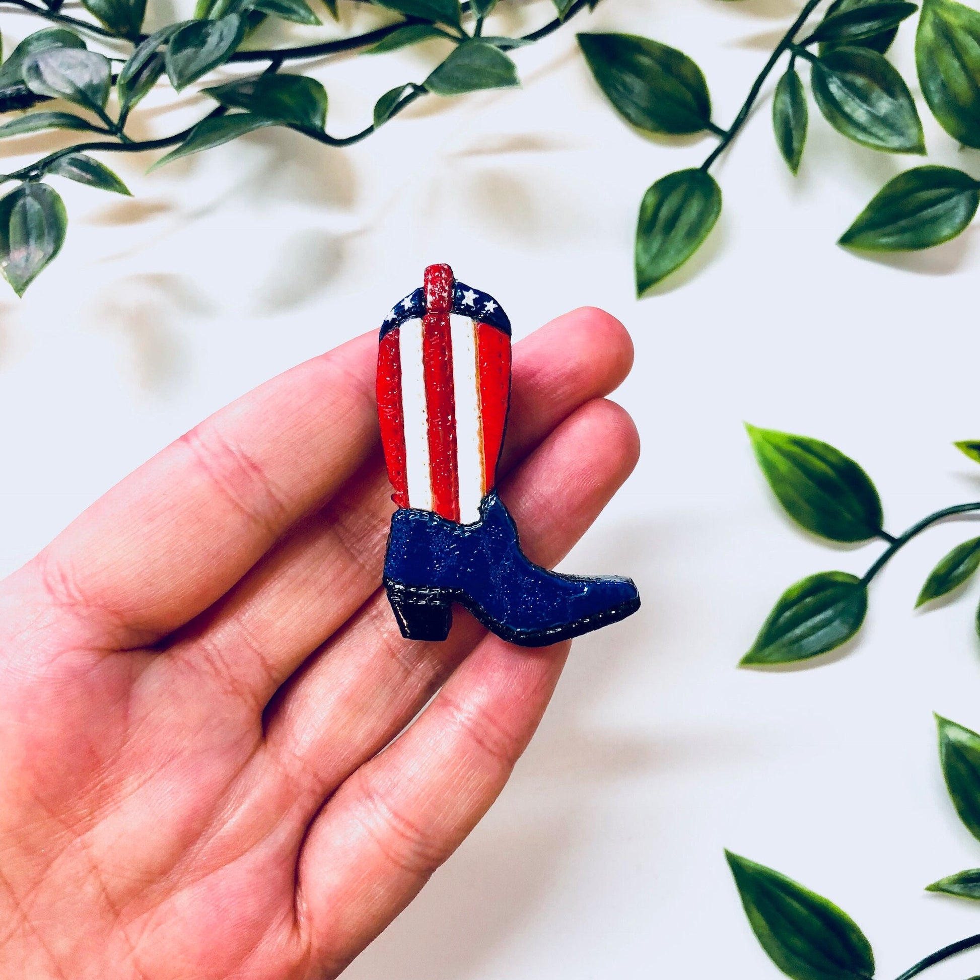 Vintage red, white and blue cowboy boot pin brooch with American flag design, held in hand against floral background.