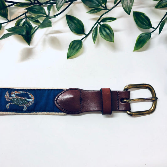 Vintage woven belt with blue crab design and brass buckle on white background with green leaves