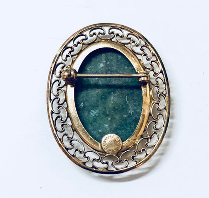 Vintage gold filled brooch with ornate scrollwork frame surrounding an oval green stone cabochon, likely jade or aventurine, against a white background.