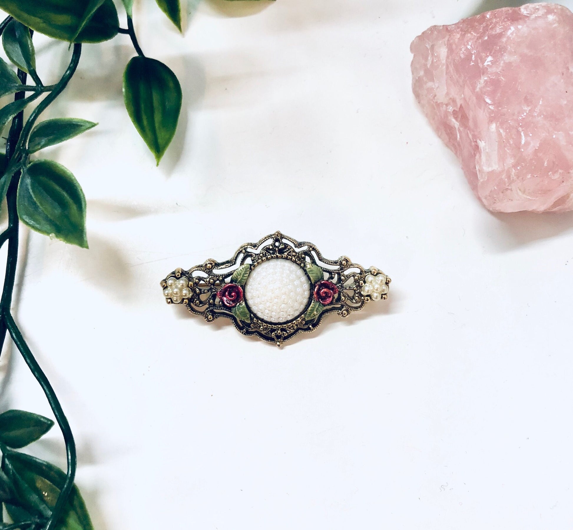 Vintage gold-toned floral brooch pin with pink rhinestones and decorative metalwork details, displayed on white fabric with a rose quartz crystal and green leafy plant.