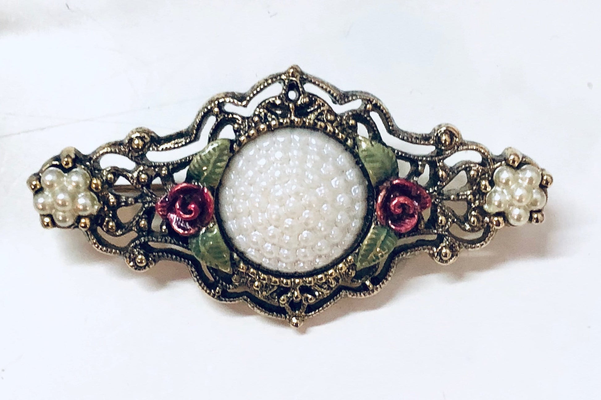 Antique gold-toned brooch with intricate floral design, featuring white beads, green enamel leaves, and pink and purple jeweled accents, set against a white background.