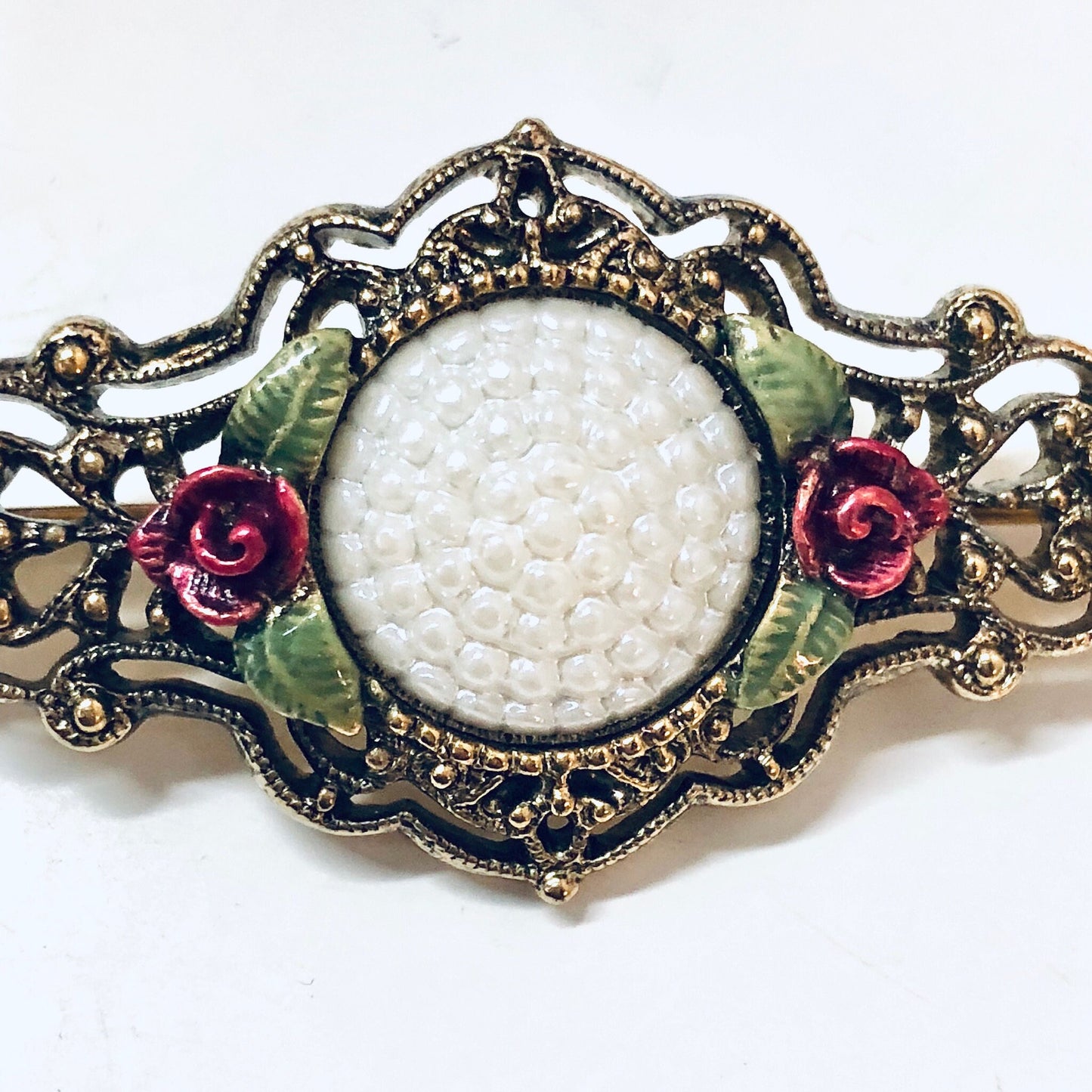 Vintage gold-toned brooch pin with intricate floral design, featuring white beads surrounded by green leaves and red rose accents, set in an ornate filigree metal frame.