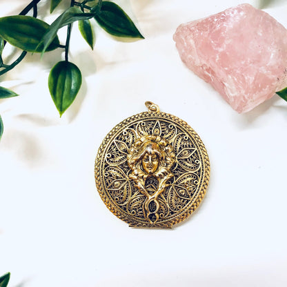 Vintage gold-toned filigree locket pendant featuring an intricate lady face design, displayed with rose quartz crystal and greenery.