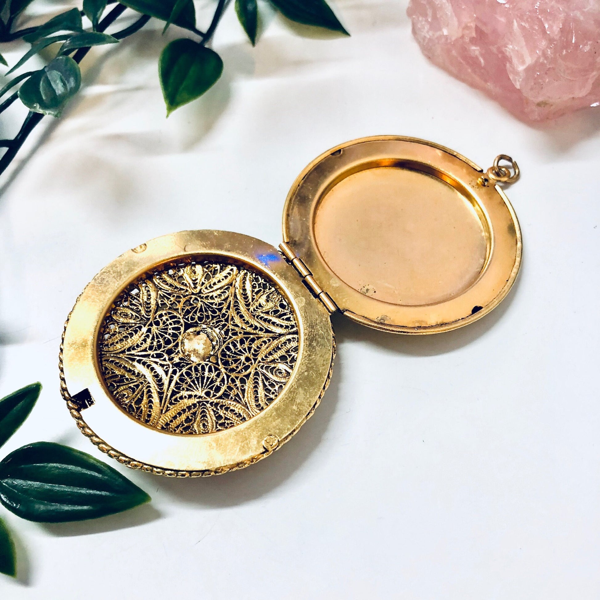 Vintage gold toned locket pendant necklace with intricate lady face design, displayed open on white marble surface with pink quartz and greenery accents.