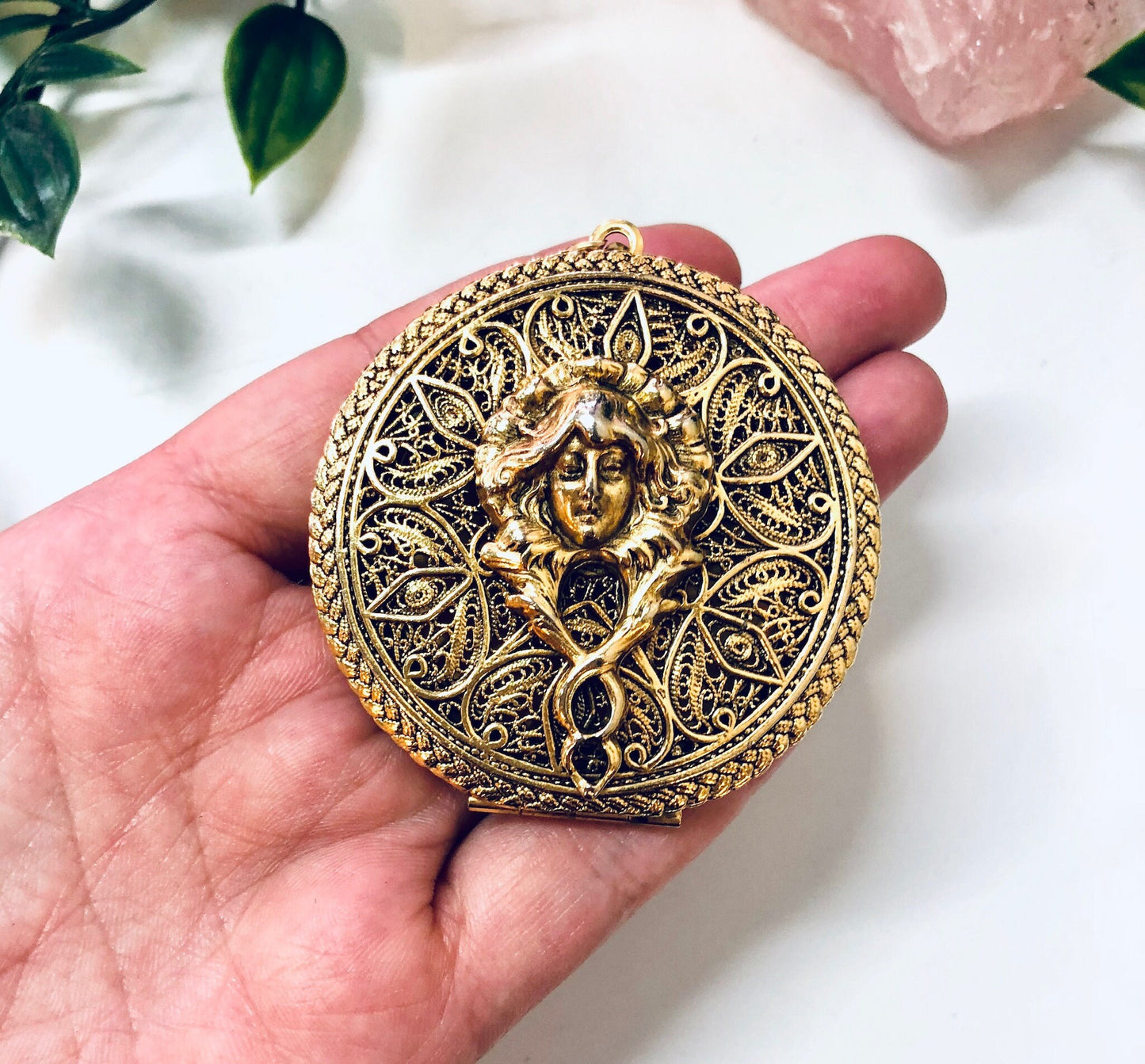 Antique gold-tone ornate round locket pendant featuring an intricate relief design of a woman's face surrounded by floral motifs, held in a person's hand with pink rose quartz crystals in the background.