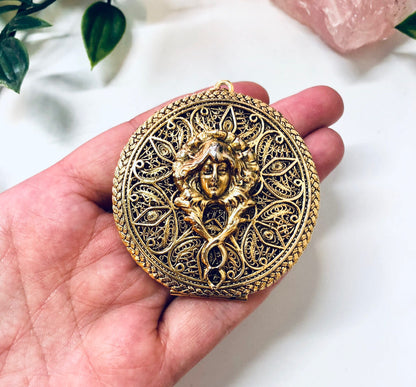 Antique gold-tone ornate round locket pendant featuring an intricate relief design of a woman's face surrounded by floral motifs, held in a person's hand with pink rose quartz crystals in the background.