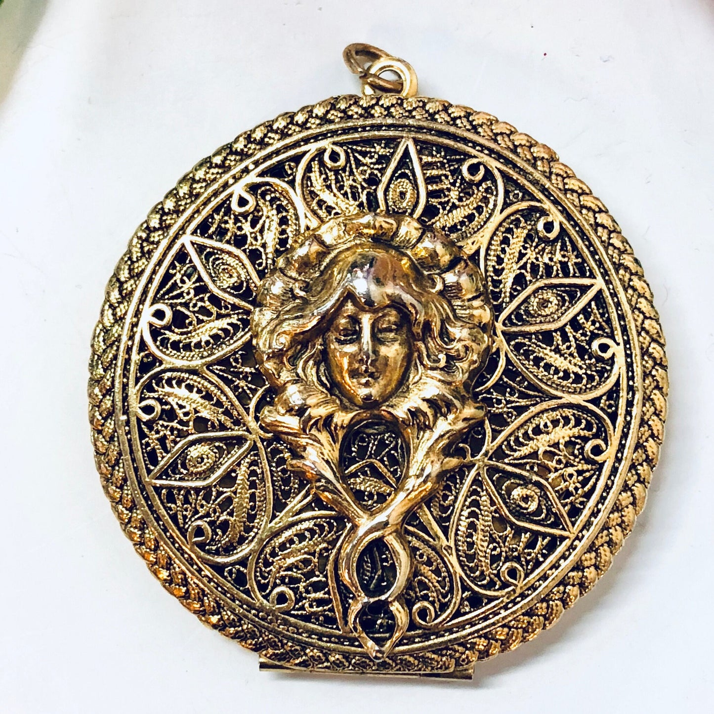 Ornate vintage gold-toned locket pendant featuring an intricate filigree design with a lady's face in the center, suitable for a necklace or other piece of jewelry.