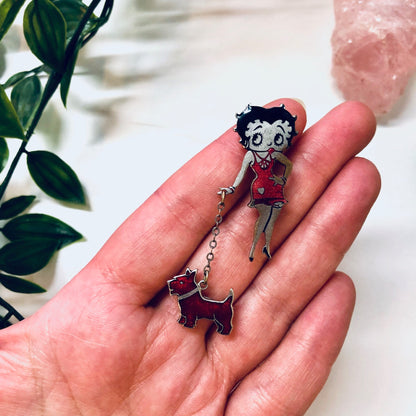 Vintage silver-tone Betty Boop pin featuring the iconic cartoon character in a red outfit holding a small black dog, set against a blurred background with greenery, showcasing retro flapper girl jewelry from a bygone era.