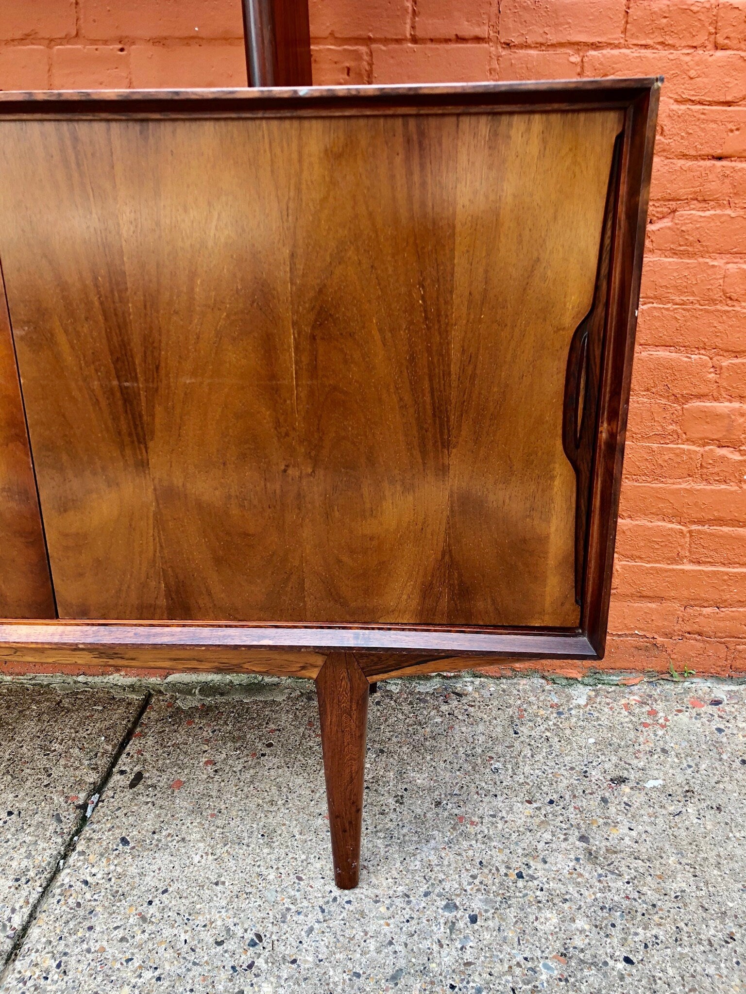 Beautiful mid-century modern teak credenza server buffet with hardwood construction and elegant curved design against a brick wall background.