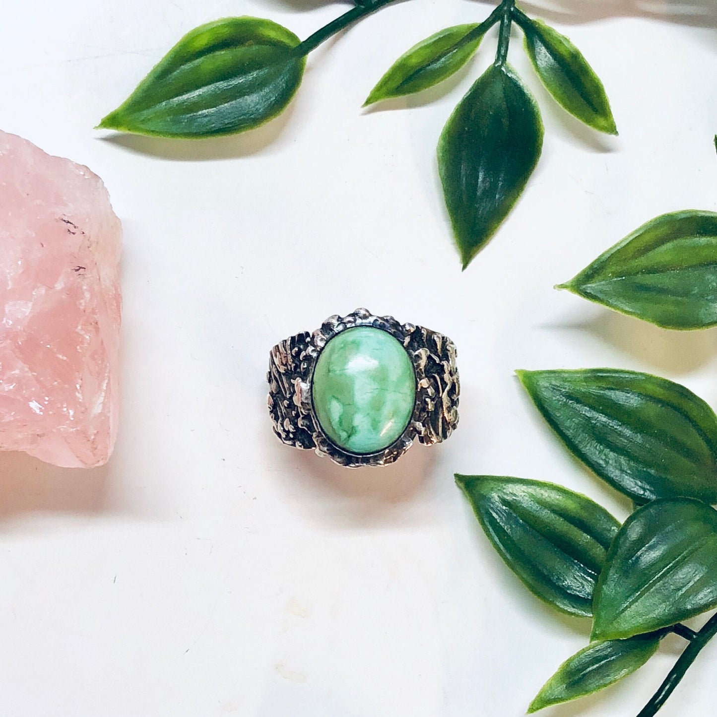 Vintage sterling silver ring with large oval turquoise stone surrounded by textured silver band, alongside rose quartz stone and green leaves, on white background