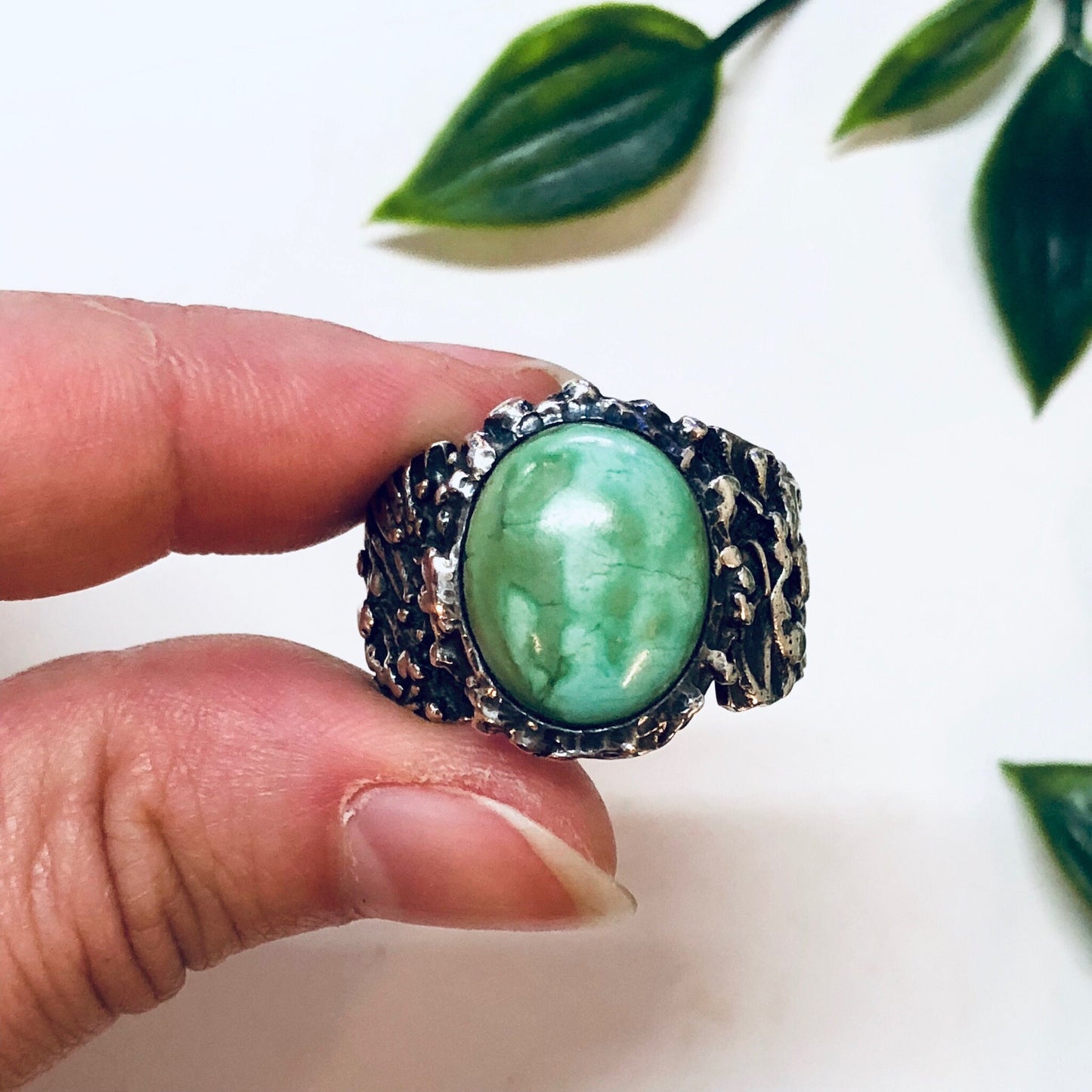 Vintage sterling silver ring with large oval turquoise cabochon set in textured silver bezel, held by a hand with green leaves in the background.