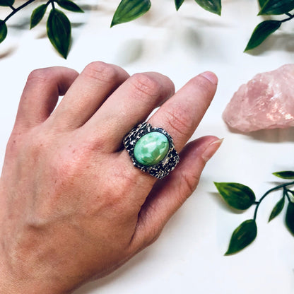 Vintage silver ring with textured band and large oval turquoise stone in shades of blue and green, shown on a hand against a white background with green leaves and rose quartz crystals.