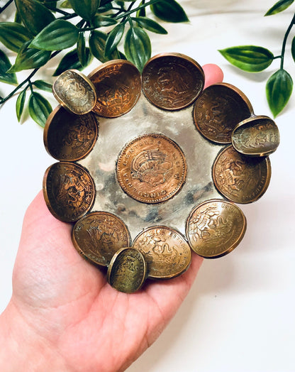 Vintage Mexican coin ashtray, ring dish or catch-all tray made from centavos copper currency arranged in a circular pattern, held in a hand with plant leaves in the background.