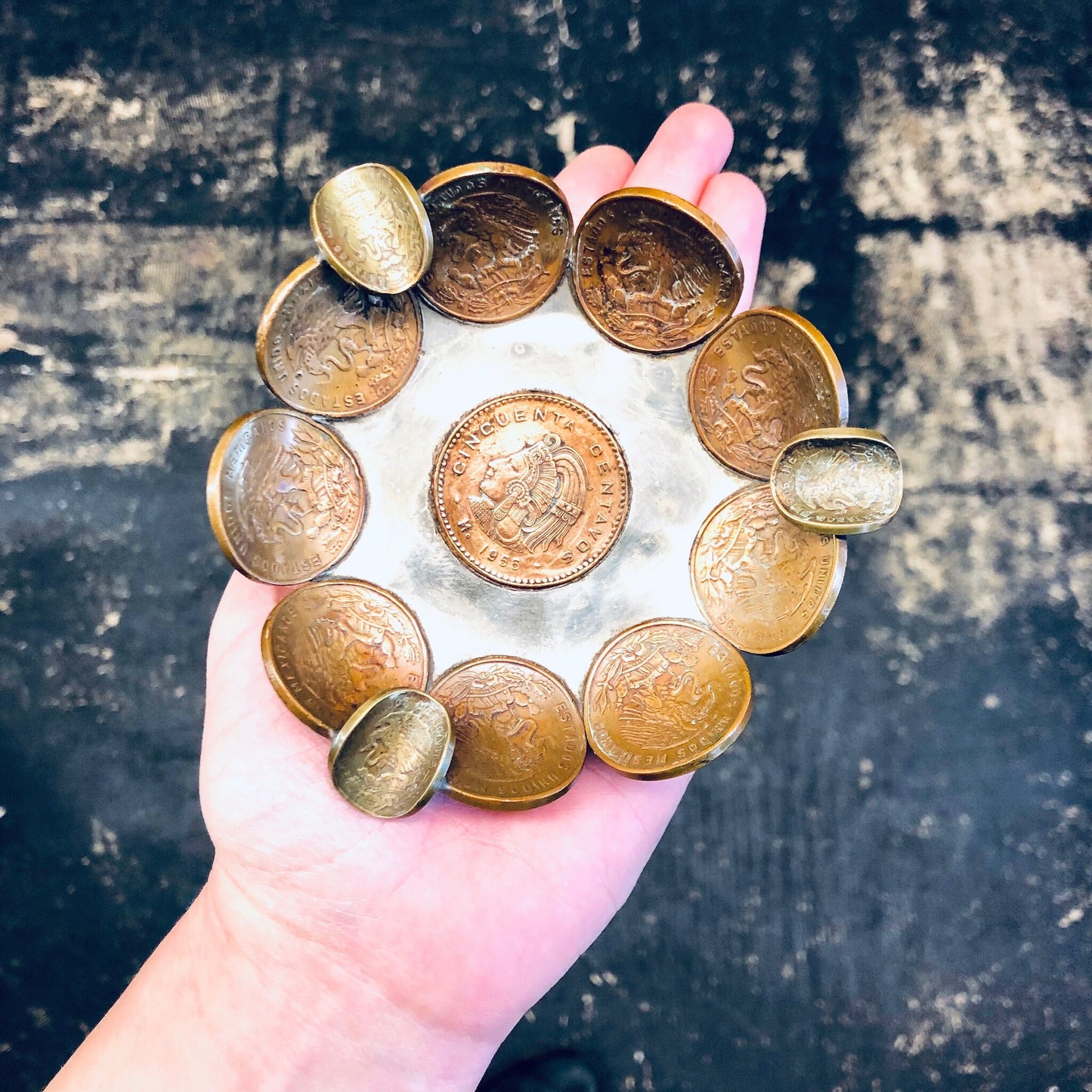 A hand holding a circular arrangement of vintage Mexican centavo coins fashioned into a decorative dish or ashtray, against a dark grungy background.