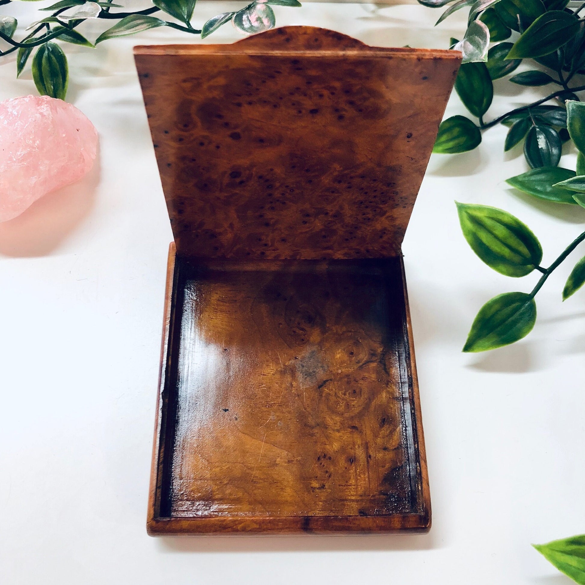Vintage wooden hinged box with burl wood grain pattern, could be used as a cigarette case, business card holder or small container, surrounded by greenery on a light colored background.