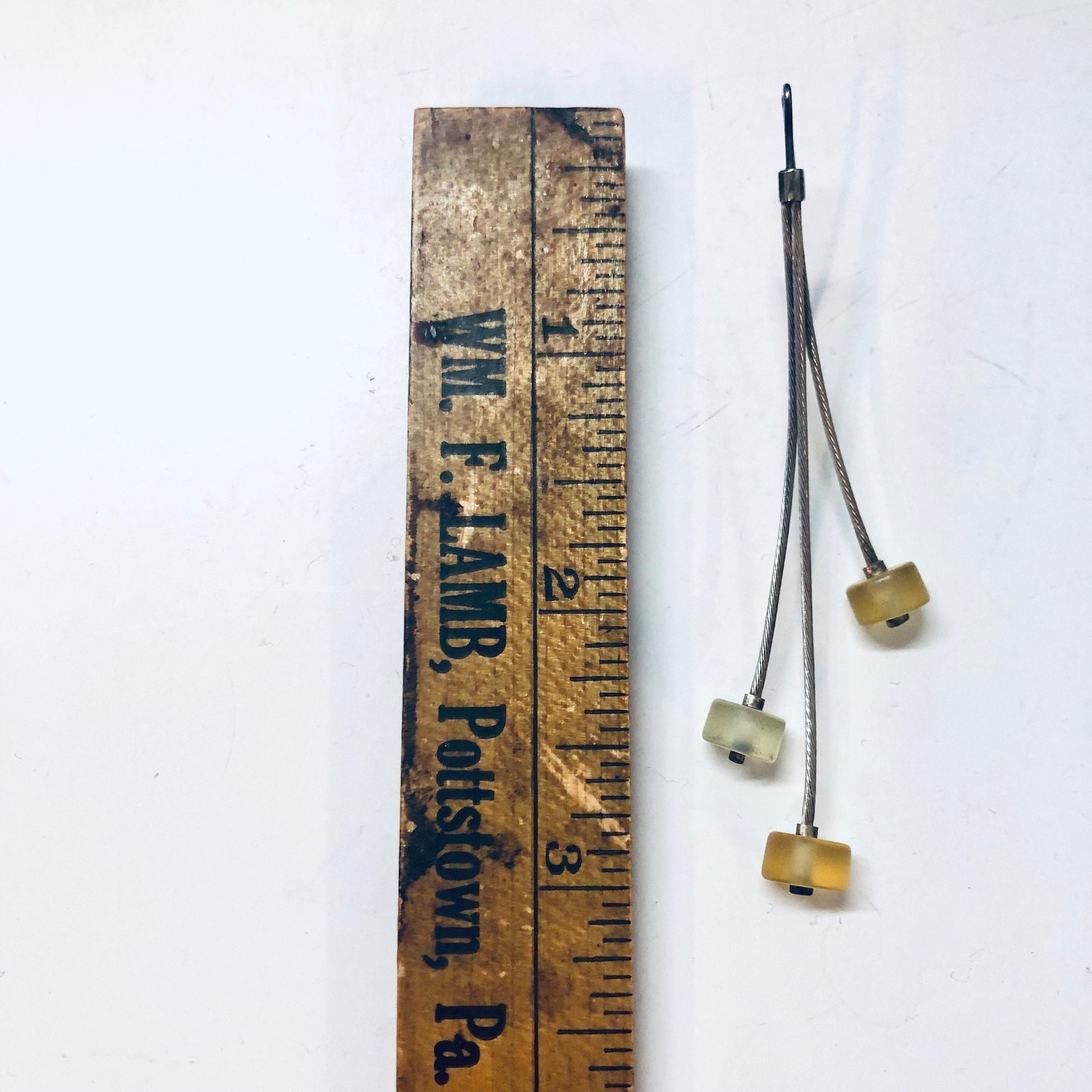 Vintage wooden ruler repurposed as earring display with three unique, long yellow and green beaded earrings hanging from a grey string against a plain white background.