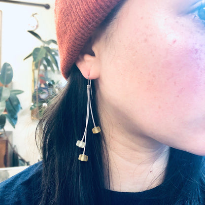 Closeup of a woman wearing long, dangly gold earrings with heart shapes at the ends. The earrings stand out against her dark hair and orange knit hat.