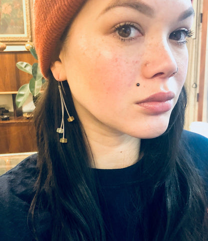 Close-up portrait of a young woman with long dark hair wearing an orange beanie hat and yellow dangling earrings, looking directly at the camera with a neutral expression.