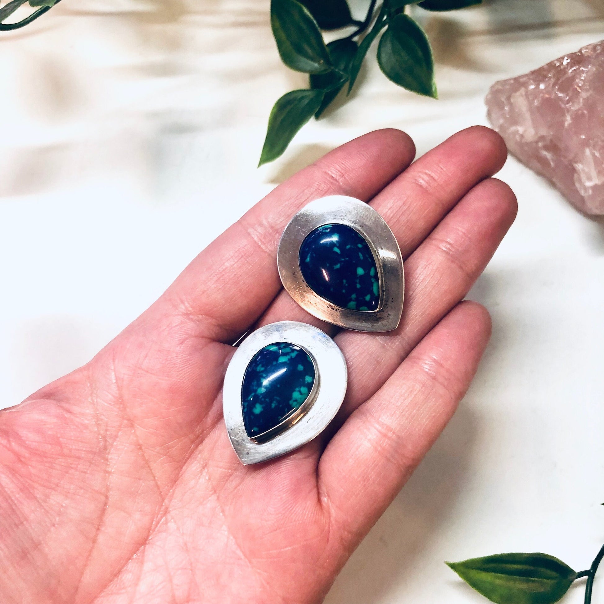 Vintage silver clip-on earrings with teardrop-shaped azurite malachite stones in shades of deep blue and green, held in open hand with leaves in background.